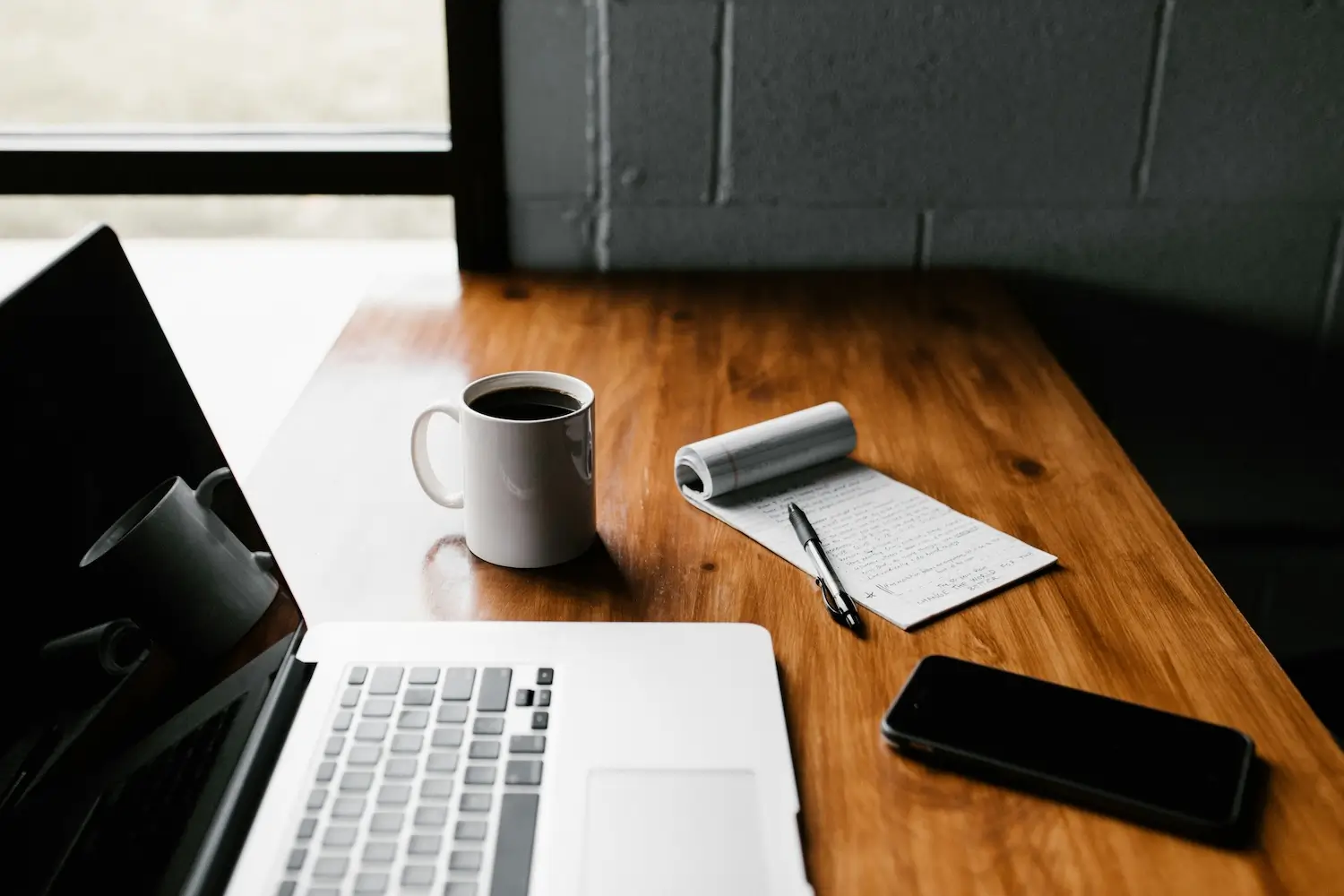 Houten bureau met een laptop, kop koffie, notitieblok met pen en een smartphone, bij een raam in een rustige werkruimte.