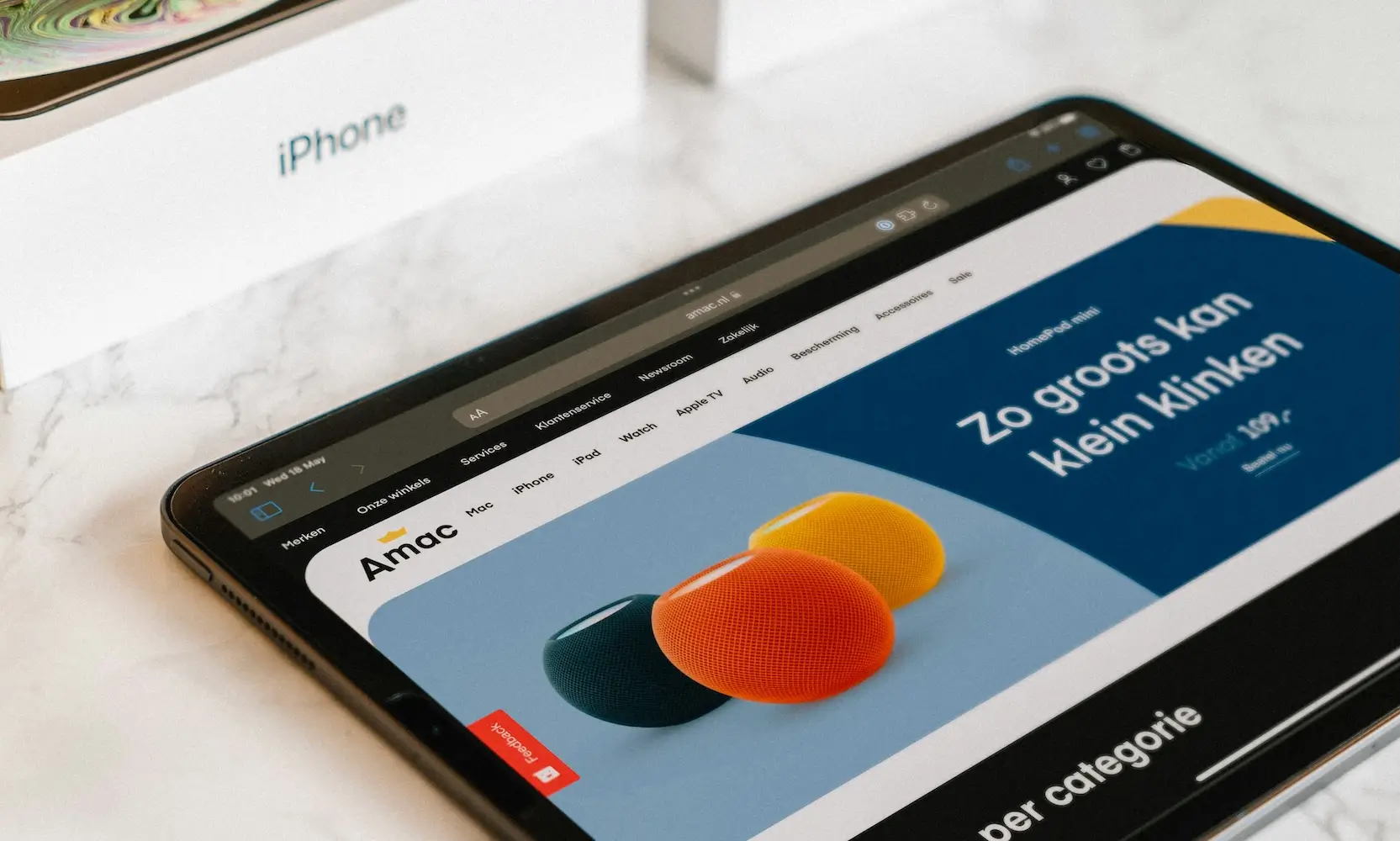 Tablet met een webshop geopend waarop kleurrijke HomePod mini’s worden getoond, liggend op een tafel met een iPhone-doos op de achtergrond.